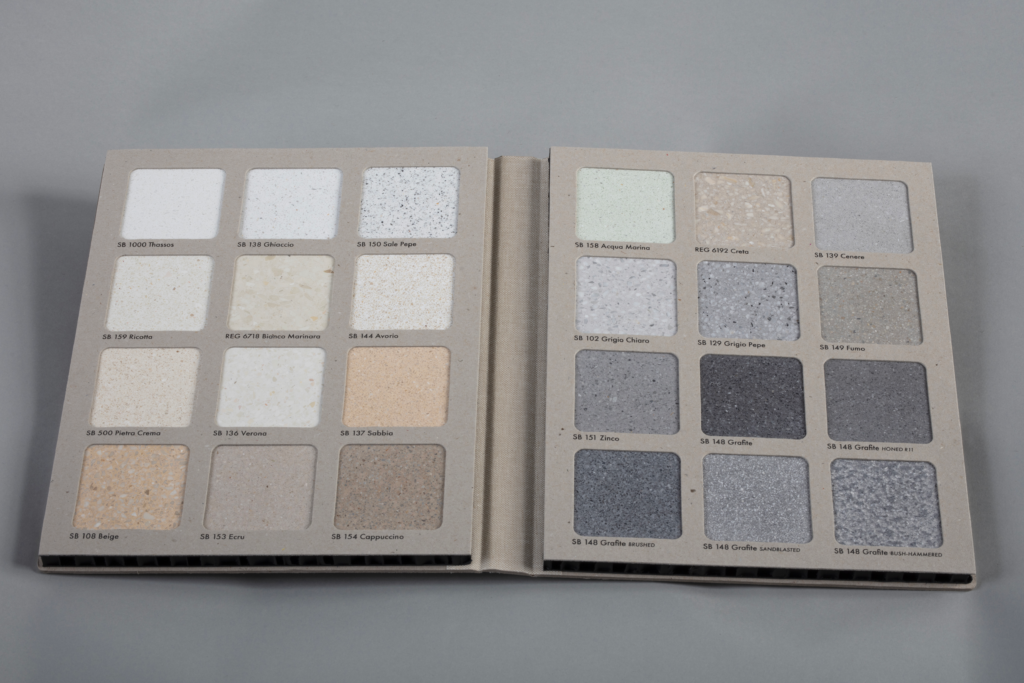 Open color swatch book showing two-page spread of square pigment samples in whites, creams, and grays with labeled names beneath each tile on a neutral beige page against a gray background.