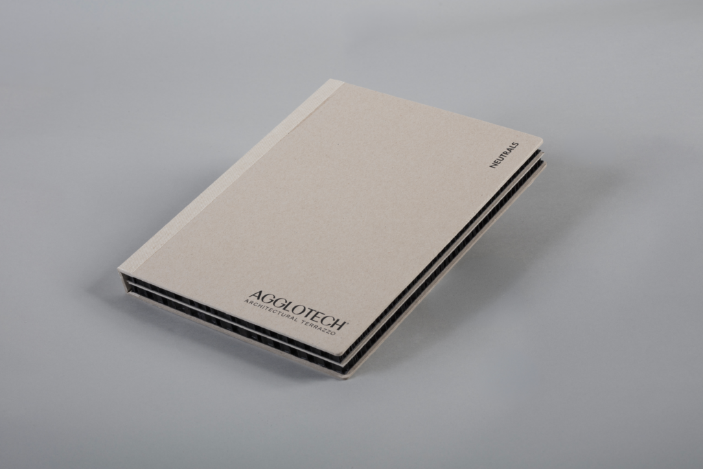 Beige architectural sample book labeled NEUTRALS, angled on a grey surface, with ribbed spine visible and AGGLOTECH Architectural Terrazzo logo near the bottom edge.
