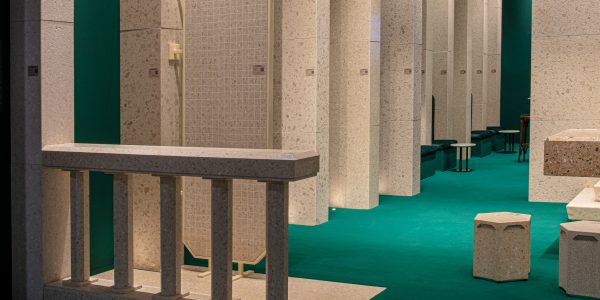 Indoor architectural space with beige terrazzo columns, green carpet, and seating along the wall; a square-tiled panel hangs in the center.
